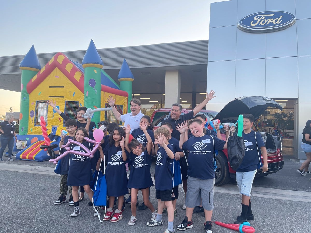 A group of kids pose for a photo in the parking lot of a Ford dealership. In the background the building with the Ford logo and a bouncy castle can be seen.